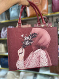 Maroon handbag with a design of a person holding flowers, held by a hand against a blurred background.