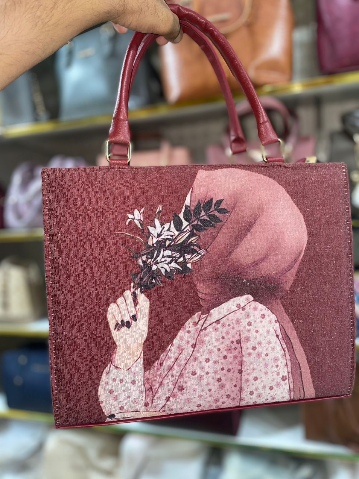 Maroon handbag with a design of a person holding flowers, held by a hand against a blurred background.